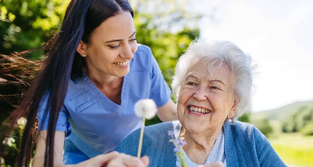 Caregiver and a smiling older woman holding a dandelion together